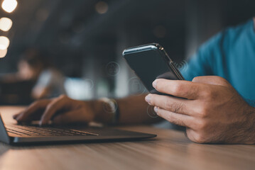 Young man's hands utilizing a laptop and checking his phone up close while working at home.