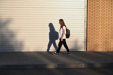 A teenage girl in black trousers and a white T-shirt walks down the street with a black backpack on her back
