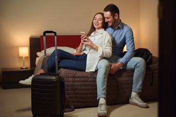 Couple browsing phone and laugh sitting on bed in hotel