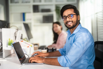 Portrait of Indian man which smiling while working with laptop at the office and looking at camera.