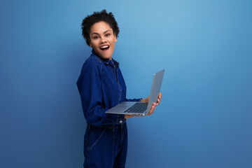 young happy brunette latin woman dressed in blue denim overalls studying remotely as a programmer and holding a laptop in her hands