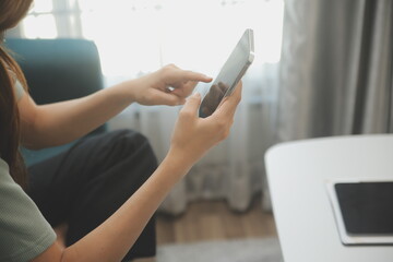 Happy Asian teen girl holding pad computer gadget using digital tablet technology sitting on the couch at home. Smiling young woman using apps, shopping online, reading news, browsing internet on sofa