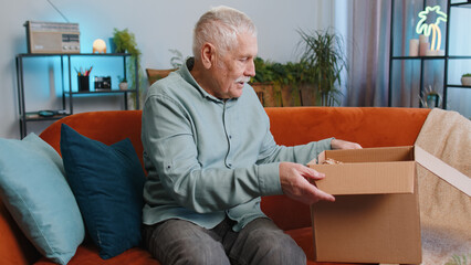 Happy senior grandfather man unpacking delivery parcel sits at home. Smiling satisfied elderly guy shopper, online shop customer opening cardboard box receiving purchase gift by fast postal shipping