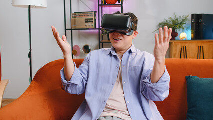 Asian man using virtual reality futuristic technology VR app headset helmet play simulation 3D 360 video game watching film movie at home apartment. Guy in goggles sits on sofa. Transition zoom effect