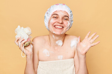 Pleased woman taking bath, holding sponge, wears shower cap and towel over body, enjoys hygienic procedures, stands with happy smile against beige background