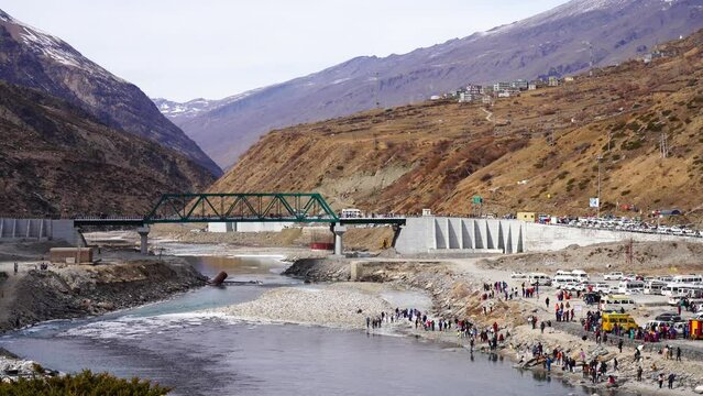 crowd of people in winter wear playing in ice on the bank of a river Chenab in lahul, manali solang with an iron bridge behind them