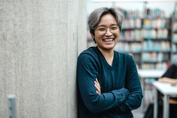 Portrait of smiling asian student standing with arms crossed in college library
