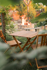 Wooden furniture in the garden against the backdrop of a fire on the patio. Among the branches of the bush stands a wooden table and chairs.