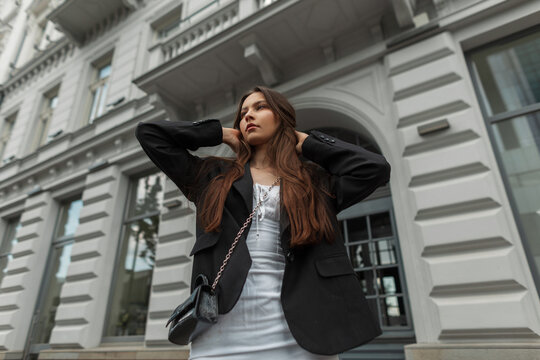 Beautiful young stylish girl model in a white dress with a black blazer with a handbag walks in the city near a vintage building