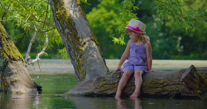 Little girl in hat and summer dress in casual clothes sitting on shore near water. Girl sitting on lake. Vacation, camping, outdoor