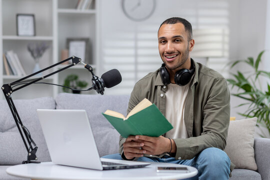Portrait of a young African American man sitting on the sofa at home in front of a laptop and a microphone and headphones with a book. Reads a lecture, records an audiobook, records a podcast