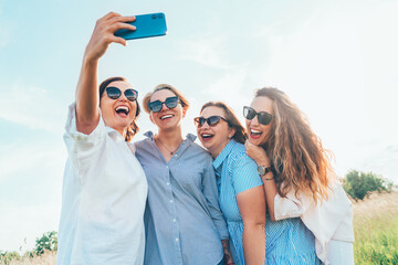 Portrait of four cheerful smiling women in sunglasses embracing together and making selfie photo using modern smartphone during outdoor walk. Woman's friendship, relations, and happiness concept image