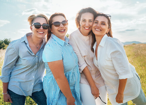 Portraits of four cheerful smiling women in sunglasses embracing and looking at camera during outdoor walking with bright sunny backlight. Woman's friendship, relations, and happiness concept image