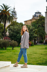 Beautiful young woman wearing jacket and jeans walking on the sidewalk. Palm trees and historic building facade on background. City recreation. Carefree person resting experiencing calmness