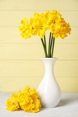 Vase with narcissus flowers on table near yellow wooden wall