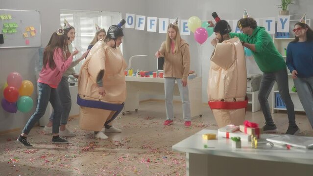 Two adult guys dressed in sumo suits wrestle jokingly. Colleagues stand around them supporting and rooting for them. Entertainment in the office. Slow motion. HDR BT2020 HLG Material.