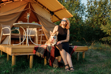 Happy young woman with Welsh Corgi Pembroke dog relaxing in glamping on summer day. Luxury camping tent for outdoor recreation and recreation. Lifestyle concept