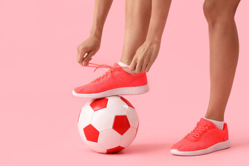 Legs of young woman with soccer ball tying shoelaces on pink background