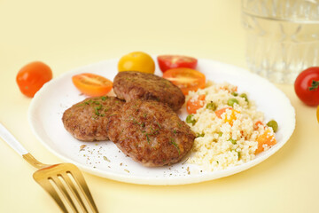 Cutlets with bulgur, boiled vegetables and tomatoes on light yellow table in kitchen