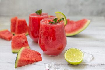Watermelon smoothies with slices of fruit and leaf of mint and lime over white wooden background.