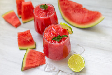 Watermelon smoothies with slices of fruit and leaf of mint and lime over white wooden background.