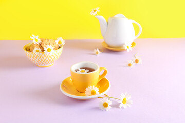 Teapot with cup of natural chamomile tea, cookies and flowers on pink table near yellow wall