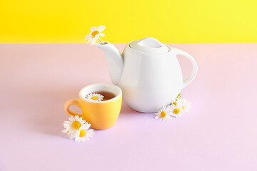 Teapot with cup of natural chamomile tea and flowers on pink table near yellow wall