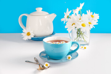 Teapot with cup of natural chamomile tea and flowers on white table near blue wall