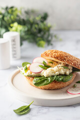 Breakfast burger with scrambled eggs, spinach, radish slices on white marble kitchen table, greenery in the background