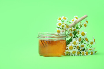 Jar of honey and fresh chamomile flowers on green background