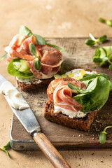 Bruschetta with prosciutto, cream cheese, cucumber, spinach and microgreens. Tasty sandwich or toasted bread on wooden rustic board. Appetizer or breakfast