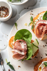 Bruschetta with ham or prosciutto, cream cheese, cucumber, spinach and microgreens. Tasty sandwich or toasted bread on white tray. Appetizer or breakfast, top view, flat lay