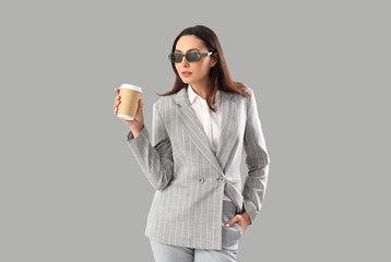 Young woman in stylish suit with cup of coffee on light background