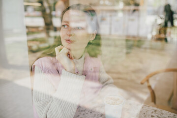 Young woman sitting near window in a coffeehouse, enjoying latte.