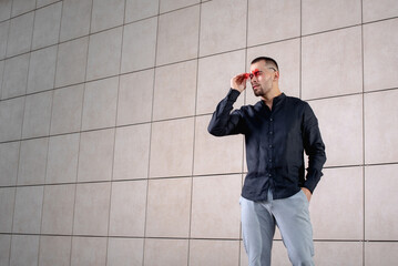 a stylish man in light pants and a black shirt against the background of an empty wall takes off his glasses and looks away angrily