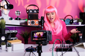Woman with pink hair recording podcast while talking with remote fans in vlogging studio, posting content on social media. Vlogger broadcasting vlog using professional filming equipment