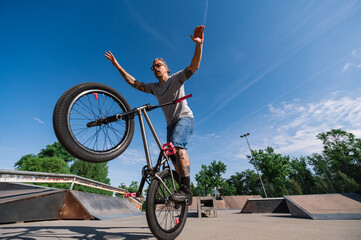 An urban mature professional bmx bike rider is performing tricks on one wheel without holding handlebars in a skate park.