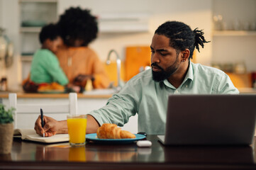 Selective focus on an african american executive working from home while his family, in a blurry background, preparing breakfast.