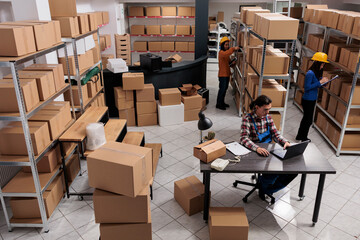 Asian postal service team doing inventory in industrial warehouse. Storehouse man and women employees checking cardboard boxes storing, managing logistics and distribution top view