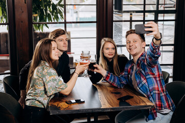 Friends cheering beer glasses on wooden table and take a selfie - People having dinner party at bar restaurant - Food and beverage lifestyle concept