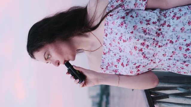 Vertical video, a happy girl in a white sundress with a floral pattern smokes a vape near the river on the embankment. A brunette in a white dress walks along the promenade
