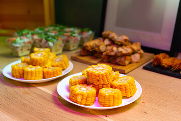 Boiled corn cut in pieces on white plates on wooden table. Table full of snacks. Selective focus.