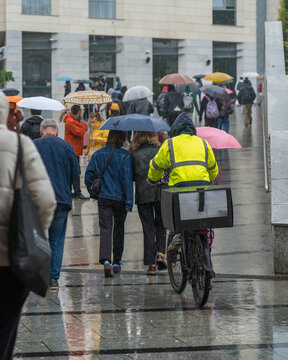 Pizza delivery man on an electric bike in the rain among real passers-by during bad weather on a city street, people with umbrellas, hoods and no umbrellas, reflections of passers-by on a wet road