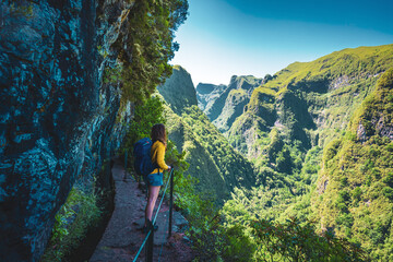 Backpacker woman enjoing view from steep cliff jungle hiking trail  next to canal through Madeiran rainforest. Levada of Caldeirão Verde, Madeira Island, Portugal, Europe. - obrazy, fototapety, plakaty