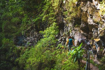 Obraz premium Tourist woman walking below big rock wall along water canal through Madeiran rainforest. Levada of Caldeirão Verde, Madeira Island, Portugal, Europe.