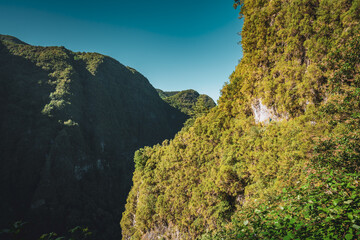 Backpacker woman in distance walking along sunny hiking trail at steep cliff through Madeira's rainforest. Levada of Caldeirão Verde, Madeira Island, Portugal, Europe.