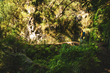 Backpacker woman walking along sunny hiking trail below large rock wall along water channel through Madeira's rainforest. Levada of Caldeirão Verde, Madeira Island, Portugal, Europe.