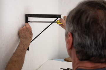 Hombre de edad mayor, atornillando una ménsula negra sobre pared blanca © AnaPliego