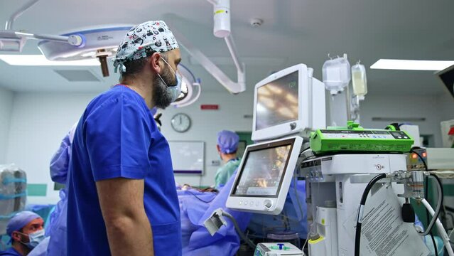 Male anesthesiologist looks attentively on the monitors showing the life signs of a patient under surgery. Doctor presses the button on the apparatus.