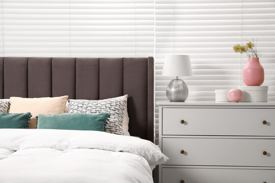 Comfortable bed with cushions, lamp and different decor on white chest of drawers in room. Stylish interior